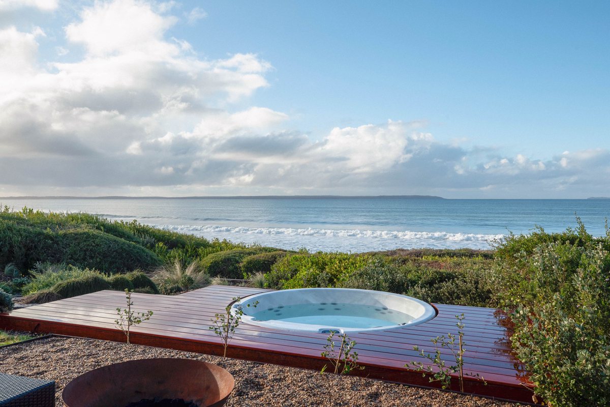 A round spa set into timber decking among coastal plants, overlooking the sea.