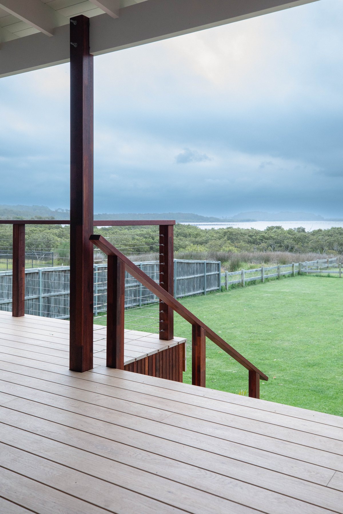 A timber deck and stair handrail beneath a roof, overlooking lawn and the sea.