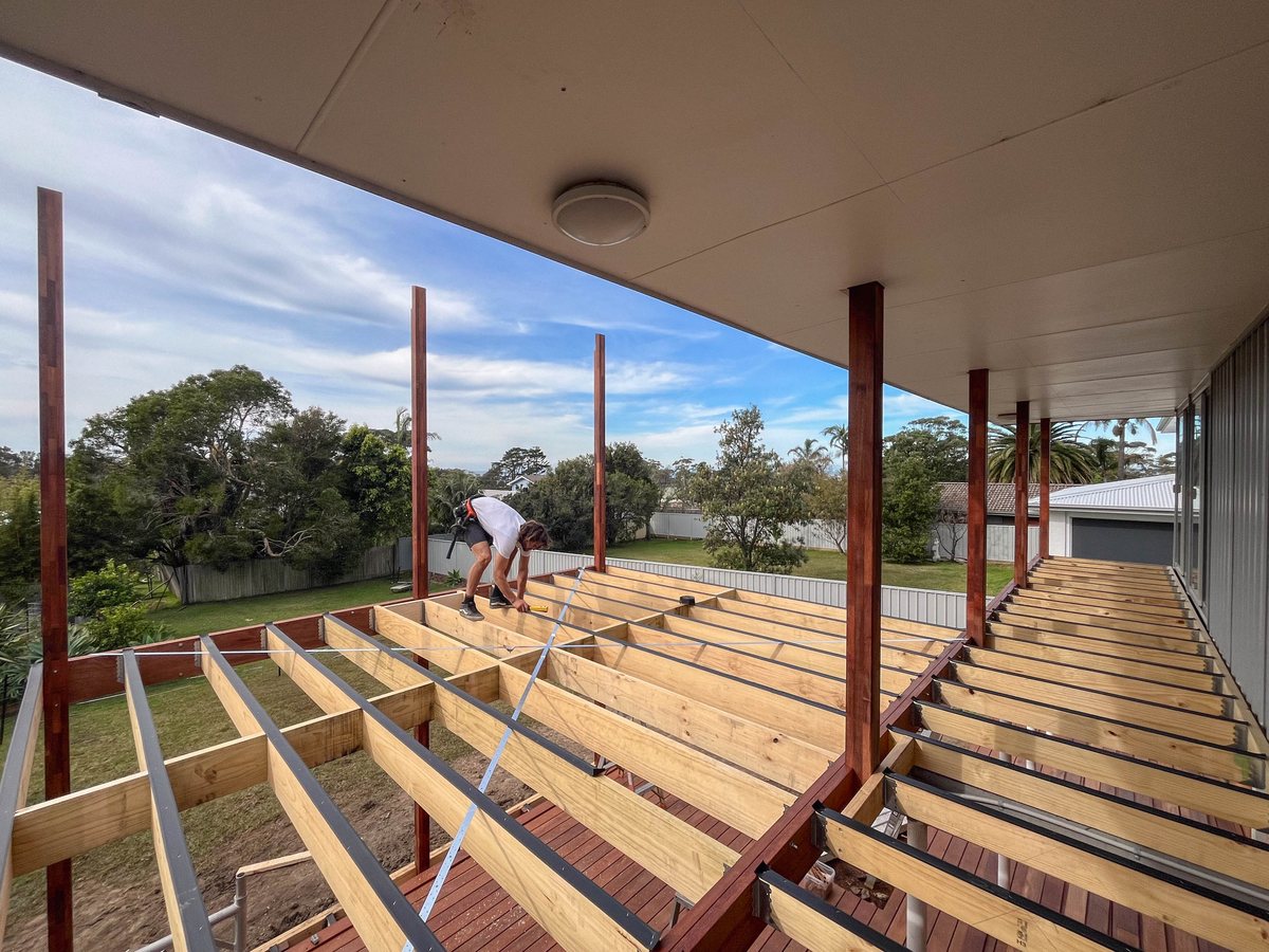 Exposed deck joists and timber posts beside a house, with a person working on the frame.