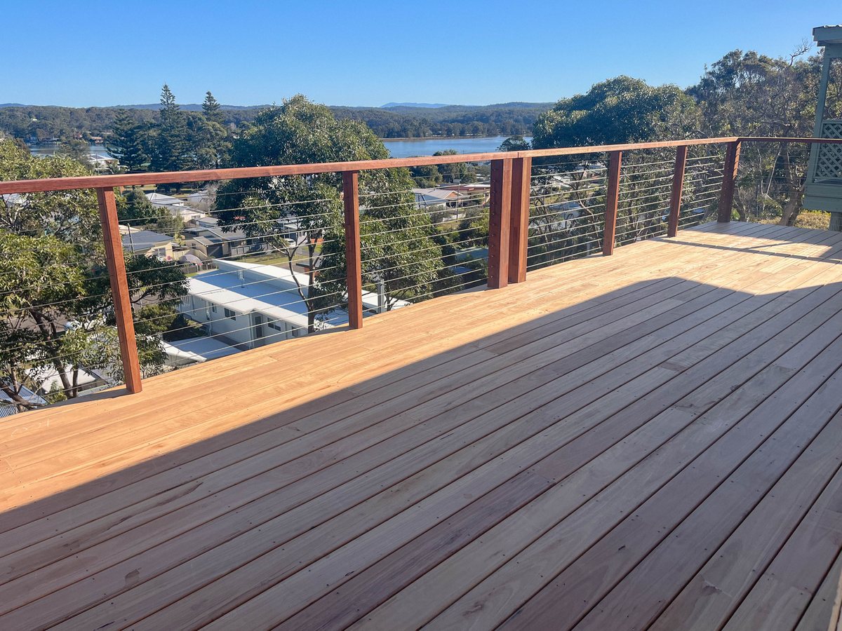 An elevated timber deck with horizontal wire balustrades overlooks houses, trees and water.