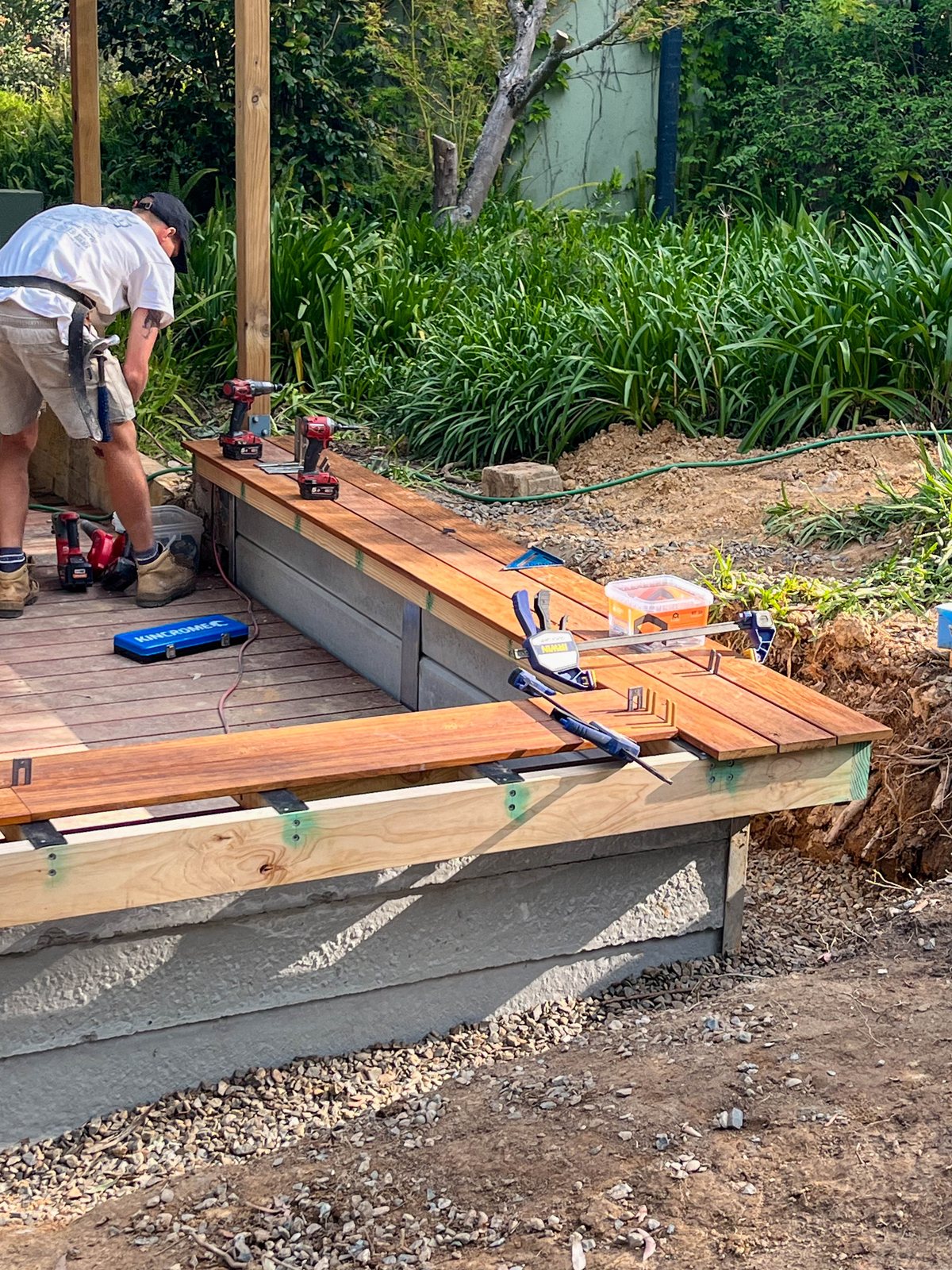A person fitting timber boards along a low wall beside a deck, with clamps and tools nearby.