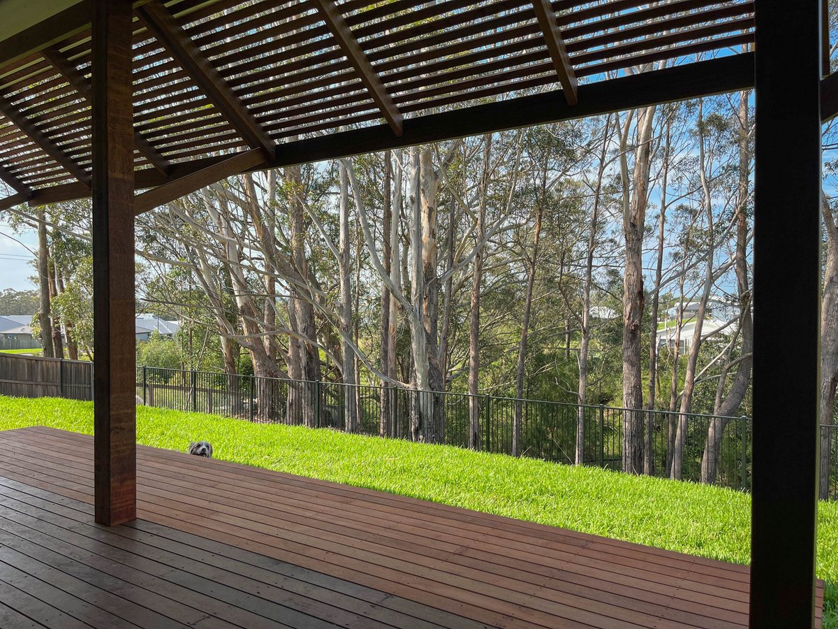 An open timber pergola with closely spaced roof slats above a deck facing lawn and trees.