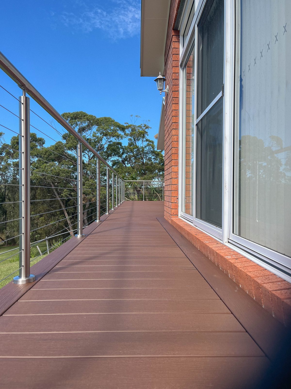 Brown decking beside a brick wall and sliding doors, with metal posts and horizontal wire balustrades.