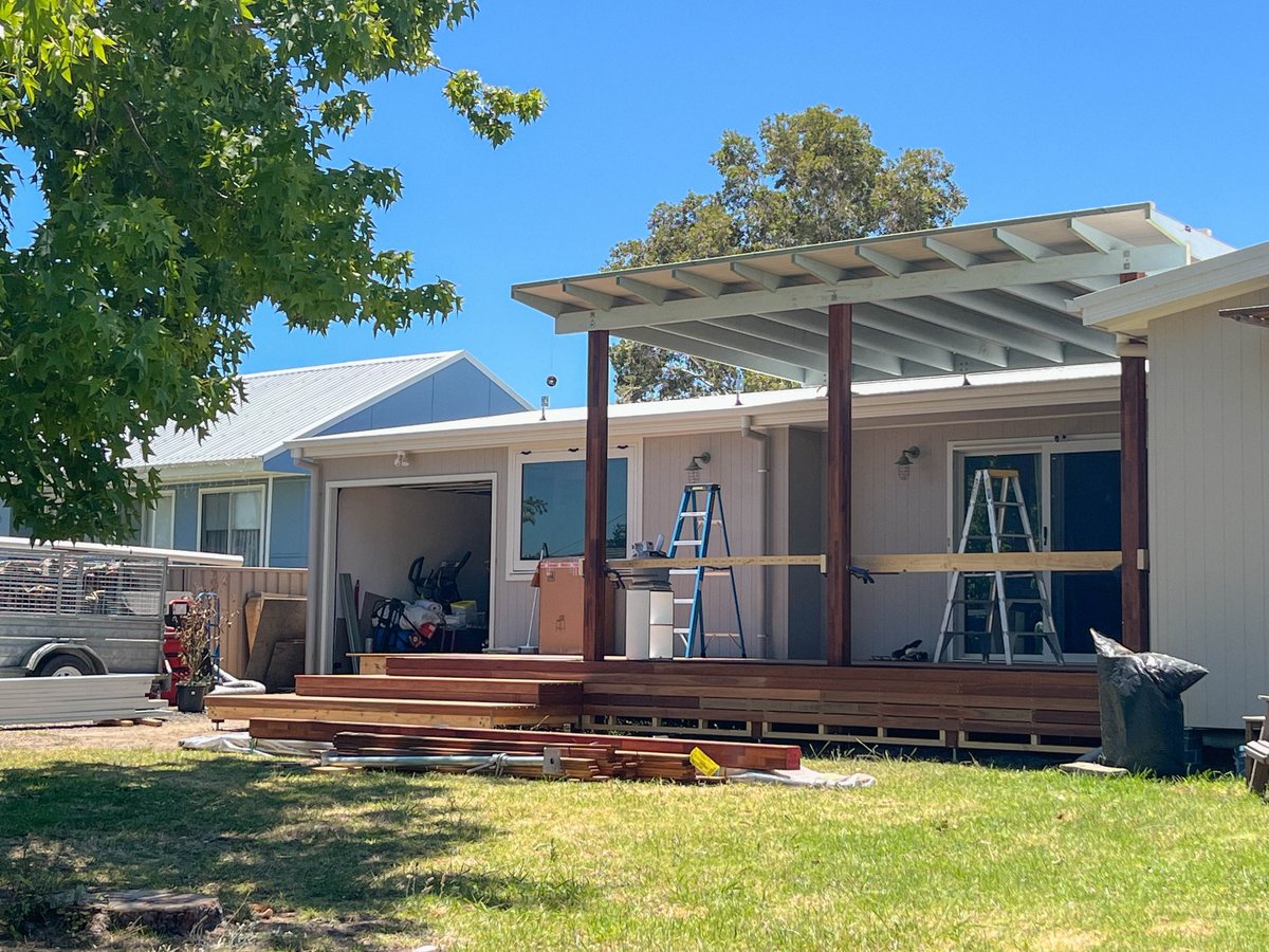 A pale single-pitch roof on timber posts above a deck, with ladders and stacked materials nearby.